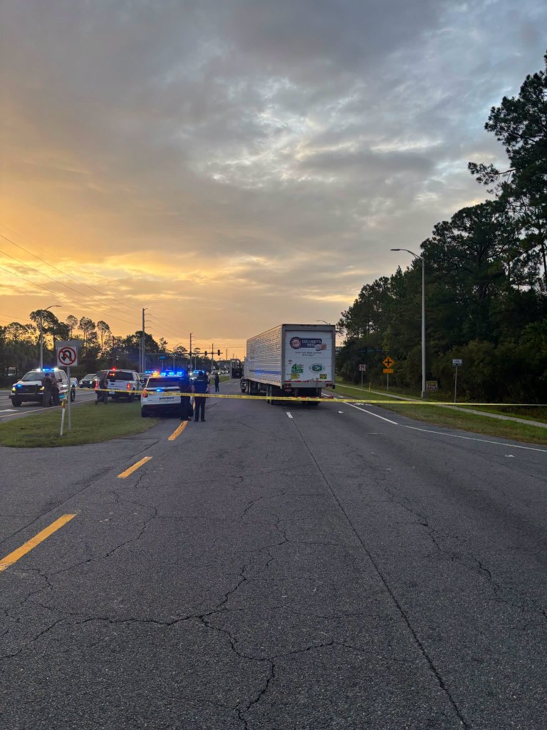 A semi-truck stopped in the middle of a highway at dusk, with police cars and officers on the shoulder and yellow police tape across the road.
