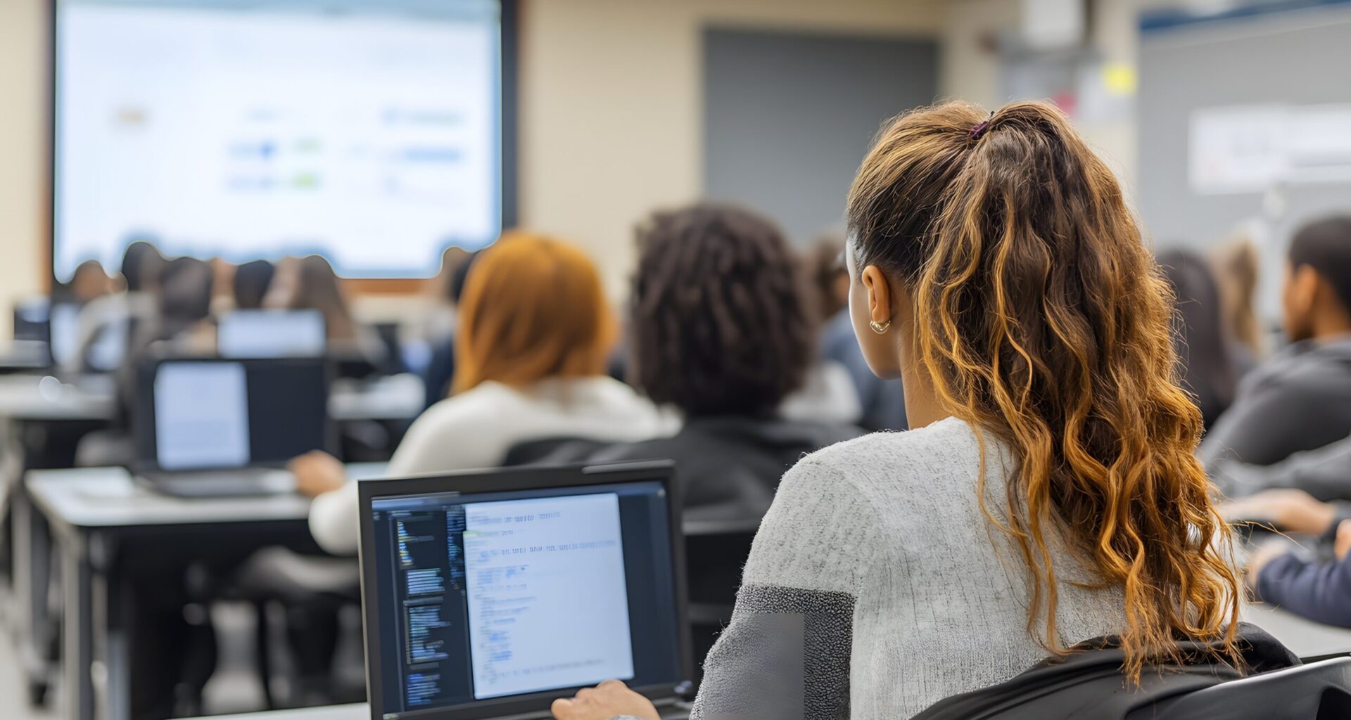 Students work on laptops in a classroom as artificial intelligence and digital learning tools are presented on a screen at the front of the room.