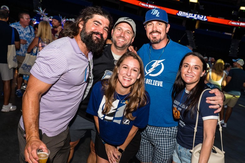 A group photograph by Nick Cardello showing five partygoers posing together at Bolts Brew Fest inside Benchmark International Arena in Tampa, Florida, on July 25, 2026. On the far left, an individual with a full dark beard and long wavy hair leans toward the camera holding a small plastic tasting cup of beer while wearing a lavender striped polo shirt. In the center foreground, an attendee with long brown hair smiles while wearing a dark blue Tampa Bay Lightning shirt, positioned in front of a person in a grey baseball cap. On the right, two attendees stand side-by-side: one wearing a blue "BELIEVE" Tampa Bay Lightning t-shirt, a blue "TBL" hat, and plaid shorts, with an arm around an individual wearing a dark navy ringer t-shirt, denim shorts, and a cream crossbody bag. Glowing arena ribbon boards and event lighting fill the background.