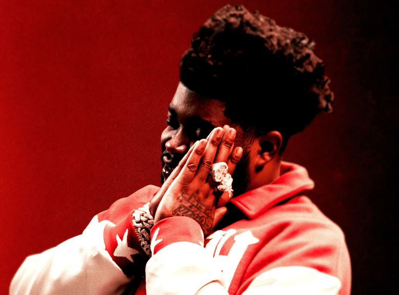 A close-up portrait cropped from the album artwork of hip-hop artist Bossman Dlow's "Dlow Curry" LP. The musician is shown in profile facing left against a solid, deep red background, with their hands clasped together near their face in a contemplative gesture. They have a short beard and dark, textured hair styled high on top, and are wearing a red and white varsity-style jacket. The artist is heavily accessorized with striking jewelry, including a thick bracelet and multiple large, sparkling rings on their fingers.