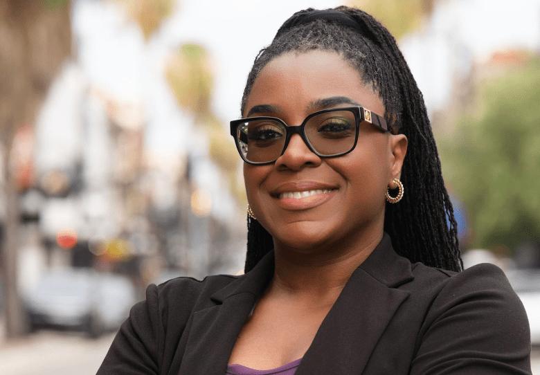 A professional campaign portrait of Tampa City Council District 3 candidate Candice Cowen smiling warmly at the camera. Framed from the chest up, the individual stands slightly turned to the side while looking forward. They have long, dark hair styled in thin micro-braids or locs gathered into a high ponytail that cascades behind them. They are wearing thick-rimmed, black cat-eye glasses, small gold hoop earrings, a purple top under a tailored black blazer, and have their arms crossed. The background features a brightly lit, softly blurred outdoor city street scene with palm tree