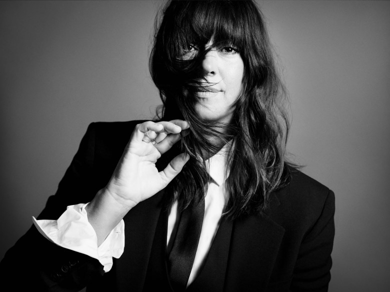 A black-and-white promotional portrait of musician Cat Power, captured from the chest up by photographers Inez and Vinoodh. The artist is dressed in a structured, dark suit jacket over a crisp white button-down shirt with a dark necktie. Their left hand is raised slightly, with the fingers gently holding a section of their long, dark hair which cascades down past their shoulders with thick, full bangs partially veiling the face. The individual looks directly at the camera with a subtle, serene expression. The portrait is set against a clean, neutral grey background with soft studio lighting.