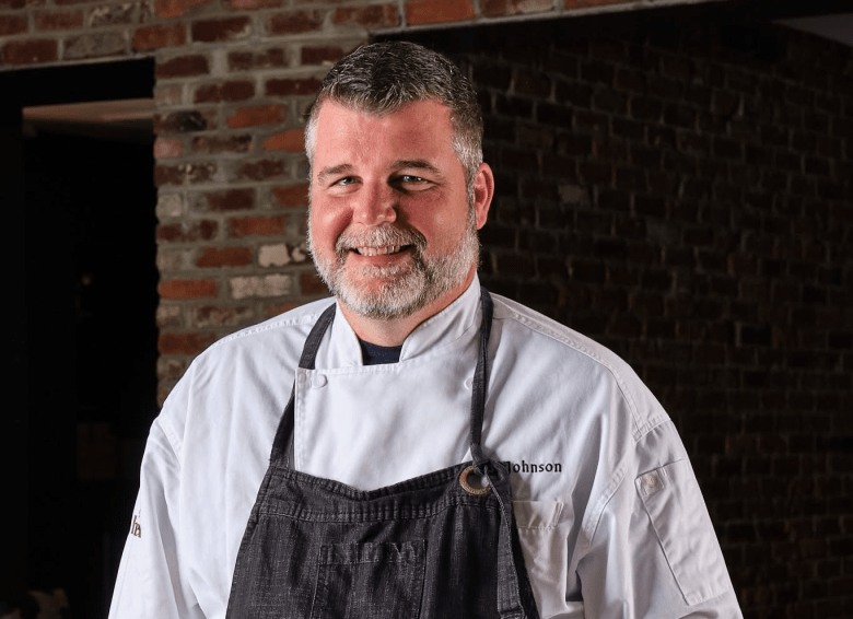 A professional portrait of Chef Chad Johnson smiling directly at the camera. The individual has short salt-and-pepper hair and a beard, and is wearing a crisp white chef's coat with the name "Johnson" embroidered on the left chest, layered under a dark grey denim apron with a metal ring on the neck strap. The background features a rustic, exposed red brick wall on the left that fades into deep, dark shadows on the right.