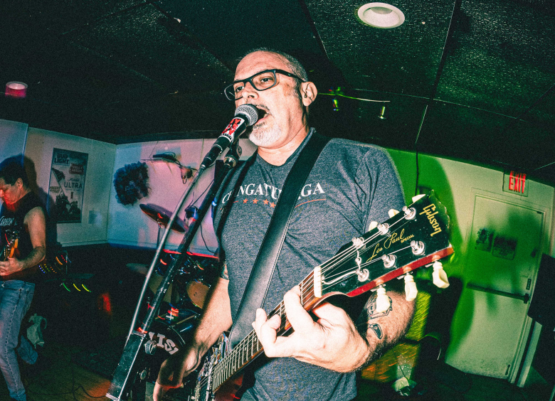 A wide-angle live music photograph of a musician from the band Chulismo performing at American Legion Seminole Post 111 in Tampa, Florida, on February 21, 2026. The artist stands in the center, singing intensely into a microphone while playing a dark Gibson Les Paul electric guitar. They have short hair, wear dark-framed glasses, and are dressed in a dark grey graphic t-shirt. To the left, another band member is partially visible playing an instrument, wearing a black sleeveless shirt and jeans. The low-lit venue features a black drop ceiling with recessed lighting, green-tinted walls with posters, and a white door illuminated by a glowing red exit sign in the background.