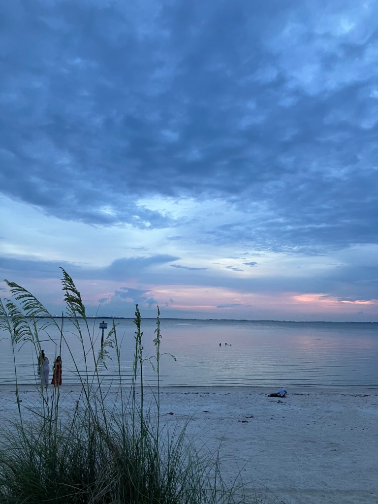 A twilight landscape photograph of Cypress Point Park beach in Tampa, Florida, featured in Creative Loafing Tampa Bay. In the foreground, tall sea grass is silhouetted against the calm water. On the left side of the white sand beach, two individuals stand near the water's edge, while another person sits on the sand further to the right. In the distance, a few swimmers wade in the water alongside a marker post, all set beneath a cloudy evening sky layered with cool blue, purple, and soft pink sunset hues at the horizon.