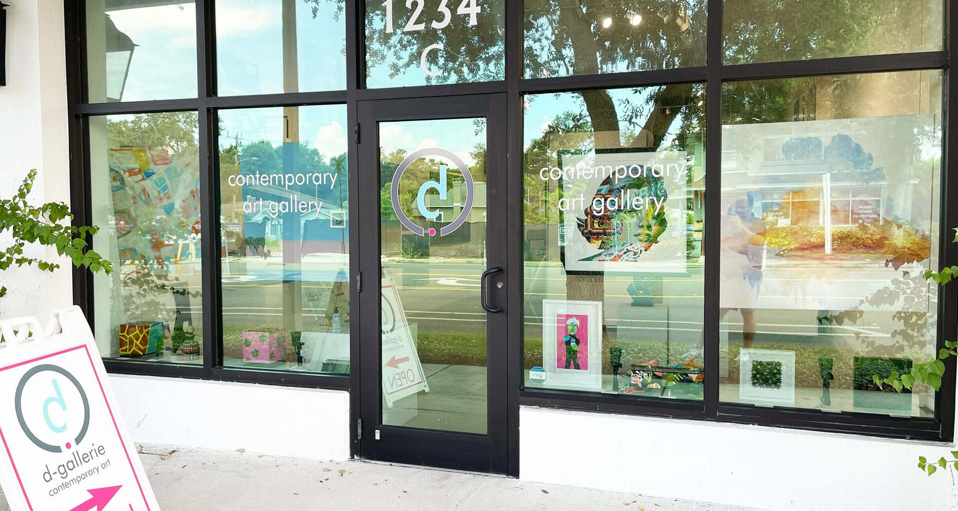 A contemporary art gallery storefront with large glass windows and a black door, displaying colorful artwork and signage with the gallery's logo and name.