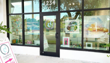 A contemporary art gallery storefront with large glass windows and a black door, displaying colorful artwork and signage with the gallery's logo and name.