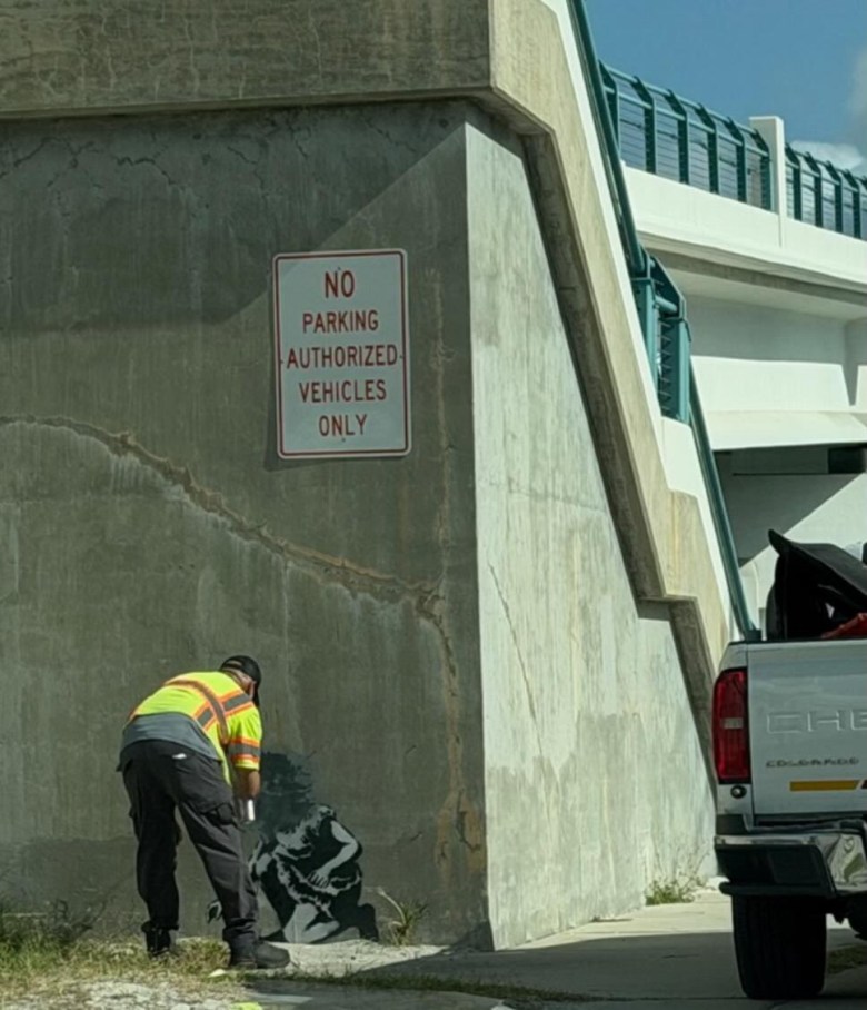 A photograph of a worker painting over stenciled street art by D.N.Z. on a concrete support wall at the John's Pass Bridge in Treasure Island, Florida, on June 23, 2026. The individual is bent down, using a spray can to cover a black-and-white stenciled figure near the base of the weathered, cracked wall. They are wearing a bright yellow high-visibility safety vest with reflective stripes, dark cargo pants, and a dark baseball cap. Mounted above them on the wall is a white sign with red text reading "NO PARKING AUTHORIZED VEHICLES ONLY." To the right, the rear tailgate and taillight of a white Chevrolet pickup truck are visible, parked along a concrete walkway under a bright, sunny sky.
