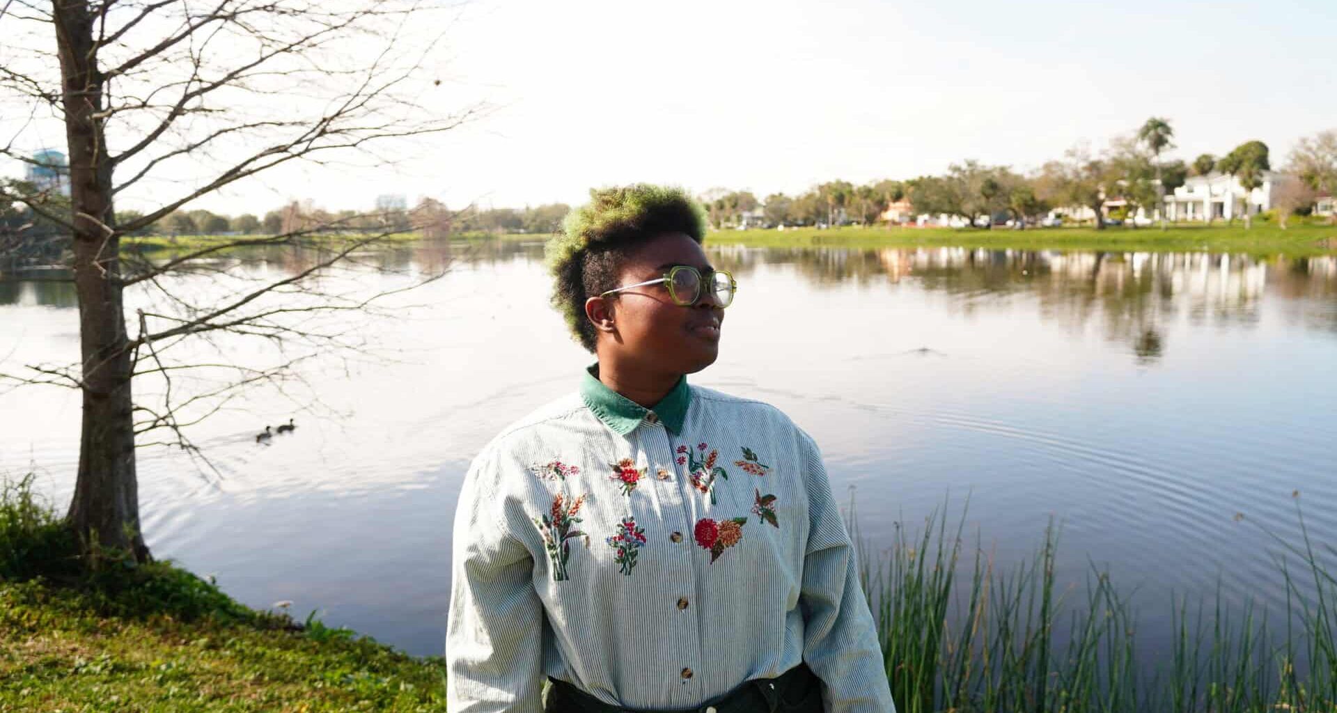 a therapist poses for a photo in a park. they are wearing a white button down shirt with flowers embroidered into it. They are standing in front of a lake on a sunny day.