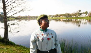 a therapist poses for a photo in a park. they are wearing a white button down shirt with flowers embroidered into it. They are standing in front of a lake on a sunny day.