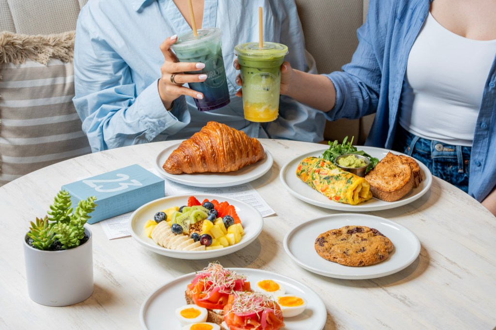 two people holding green lattes sitting at a white table covered in different prepared breakfast dishes
