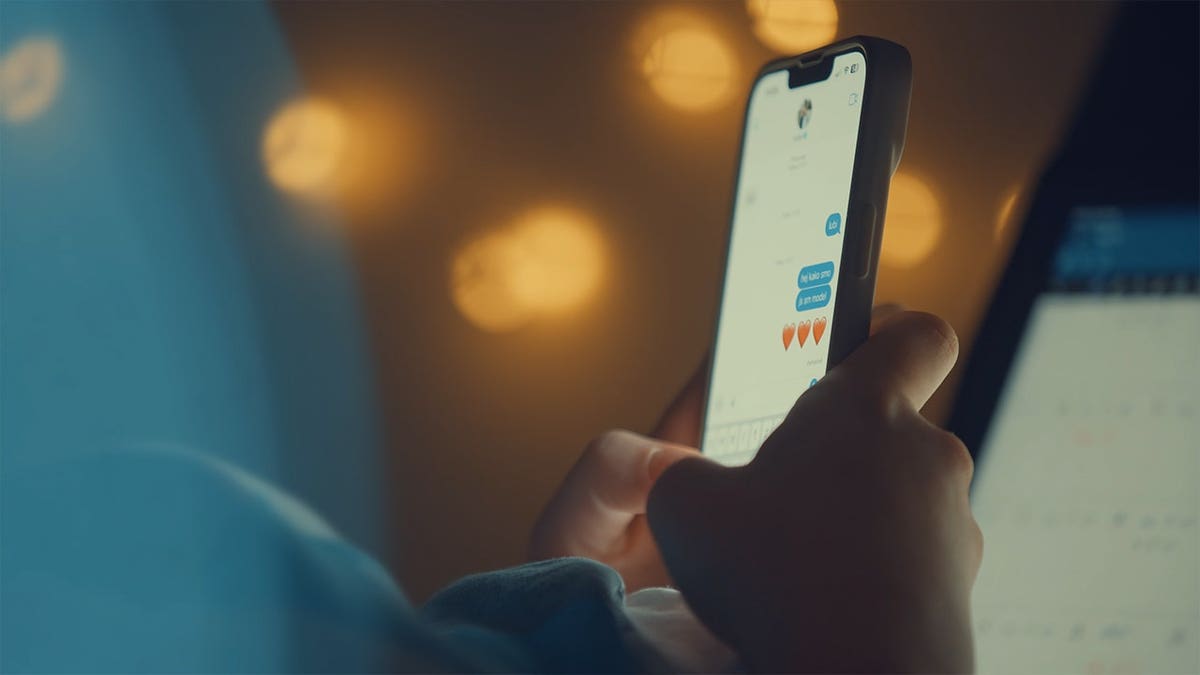 Hands of anonymous woman browsing social media app on smartphone and typing heart emoji at night, surrounded by soft bokeh of urban lights. Fingers move on screen as warm city glow contrasts with cool device light. Subtle reflections highlight hands, capturing quiet moment of digital interaction in nighttime setting.