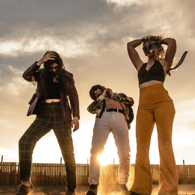 A promotional outdoor photograph of the band Deadborns featuring Jasmin Deja, Drew Giordano, and Lindsay Martin posed at sunset. The three band members stand outdoors in a row before a low wooden fence under a dramatic, cloudy sky illuminated by warm, golden sunlight low on the horizon. On the left, Jasmin Deja strikes a dynamic pose wearing a dark leather jacket, dark cropped top, and high-waisted plaid trousers, with one hand running through long dark hair. In the center, Drew Giordano stands framed by the setting sun, wearing an open patterned short-sleeve shirt over a dark chest, light-colored pants, sunglasses, and a black hat while leaning slightly backward. On the right, Lindsay Martin poses with hands behind her head, wearing a dark patterned halter crop top, round sunglasses, and high-waisted mustard-yellow flared trousers.