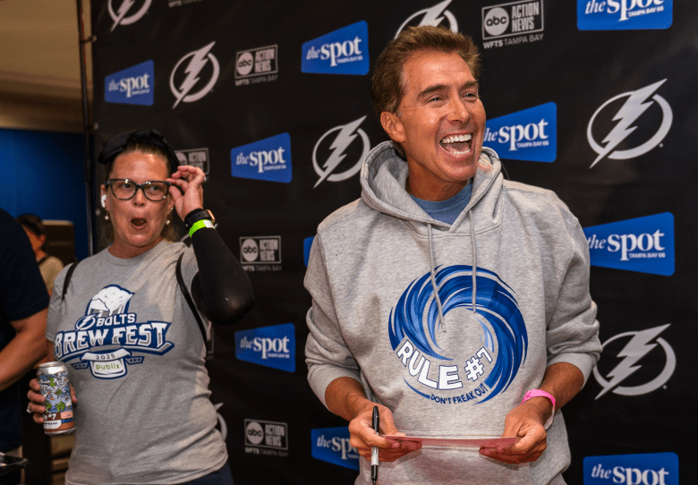 A photograph by Nick Cardello showing meteorologist Denis Phillips interacting with a fan at the Bolts Brew Fest at Amalie Arena in Tampa, Florida, on August 1, 2026. On the right, Denis Phillips laughs heartily while holding a marker and a red lanyard card, dressed in a grey heathered hoodie bearing a blue hurricane swirl graphic with the text "RULE #7 DON'T FREAK OUT." On the left, an individual wearing a grey "BOLTS BREW FEST" t-shirt, black arm sleeve, and black-rimmed glasses holds a beverage can with an wide-eyed expression while adjusting their glasses, set against a dark backdrop branded with Tampa Bay Lightning hockey logos and news sponsor logos.