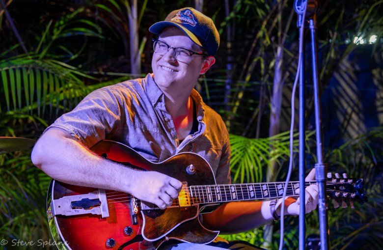 A photograph by Steve Splane showing jazz guitarist Dominic Walker performing live outdoors at DI Coffee Bar on Davis Islands in Tampa, Florida, on June 7, 2024. Positioned center frame in a medium shot, Walker smiles warmly while playing a sunburst hollowbody electric-acoustic guitar. He wears a short-sleeve printed button-down shirt over a white undershirt, glasses, and a dark baseball cap, set against a background of lush green tropical foliage under dramatic evening stage lighting with microphone stands in the foreground.