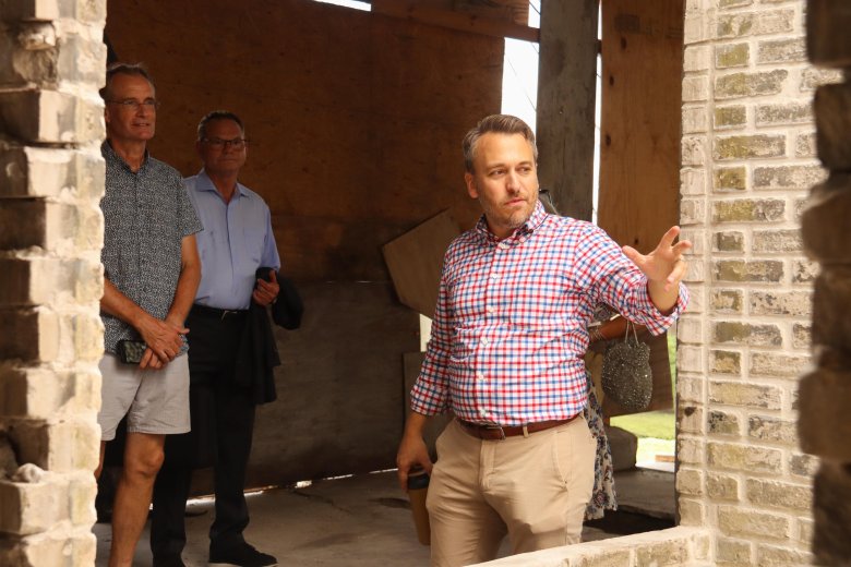 A photograph of fourth-generation J.C. Newman Cigar Co. owner Drew Newman leading an Archives Awareness Week tour inside the Sanchez y Haya building in Tampa, Florida, on July 15, 2025. In the foreground, the guide, wearing a red, white, and blue checkered button-down shirt and khaki pants, gestures outward with one hand while holding a dark tumbler in the other. In the background, two attendees stand listening closely. One individual wears glasses, a patterned blue short-sleeve shirt, and light grey shorts while holding a smartphone. Beside them, another person wears glasses and a light blue button-down shirt while holding a dark overcoat. The unfinished interior of the building appears to be under renovation, featuring a rugged exposed brick wall on the far left and raw plywood panels in the background.