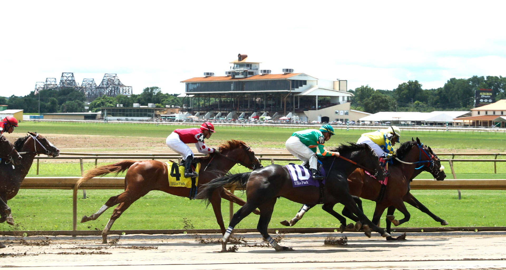 A group of racehorses gallop with their jockeys around a bend in the track. The Grandstand of Ellis Park frames the backdrop of the photo. (Photo: ©Coady Media)