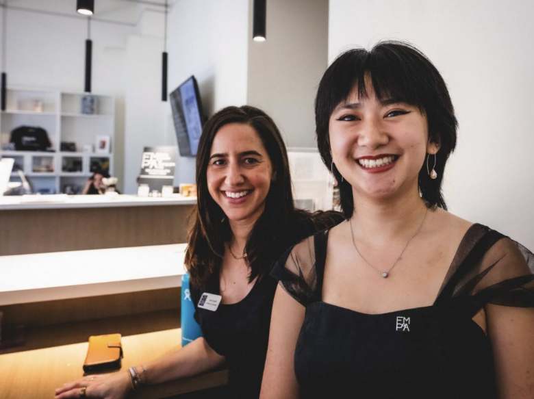 A portrait photo of two individuals at the front desk of the Florida Museum of Photographic Arts (FMoPA) in Ybor City, Florida. Positioned in the foreground on the right, a person with dark, chin-length hair with bangs, silver drop earrings, and a dark dress with sheer black tulle straps smiles warmly at the camera while wearing a small square pin reading "FM PA." To their left, standing behind the light-colored wooden reception desk, a person with long dark hair smiles as well, dressed in a black sleeveless top with a blue name tag pinned to the strap. The modern, brightly lit interior features high white walls, hanging black cylinder pendant lights, and display shelves with photography equipment and materials in the background.