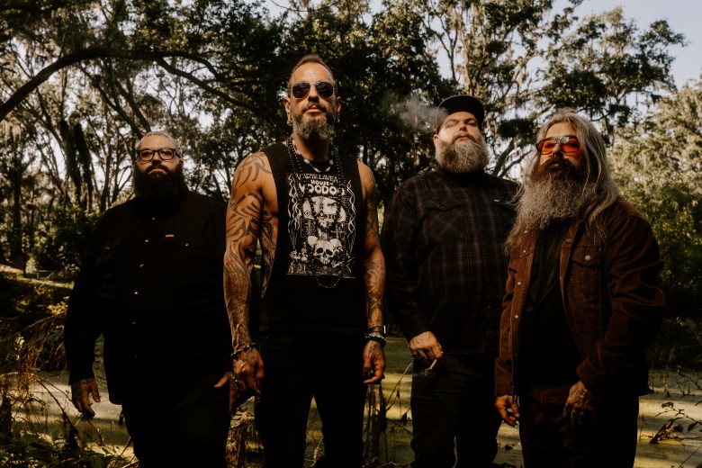 A promotional photograph by Evan Dell of the Tampa-based stoner psych-rock band Florist standing in a sunny, wooded swamp setting. Captured from a straight-on angle, the four band members stand side-by-side outdoors among trees and lush vegetation. From left to right: an individual wearing round glasses, a black button-down shirt, and black pants; a person with extensive sleeve tattoos, sunglasses, and a black graphic tank top with one hand resting on their hip; an individual wearing a dark plaid button-down shirt and a black baseball cap with puffing smoke nearby; and a person with long grey hair, a full long beard, tinted aviator sunglasses, and a brown jacket over a dark shirt.