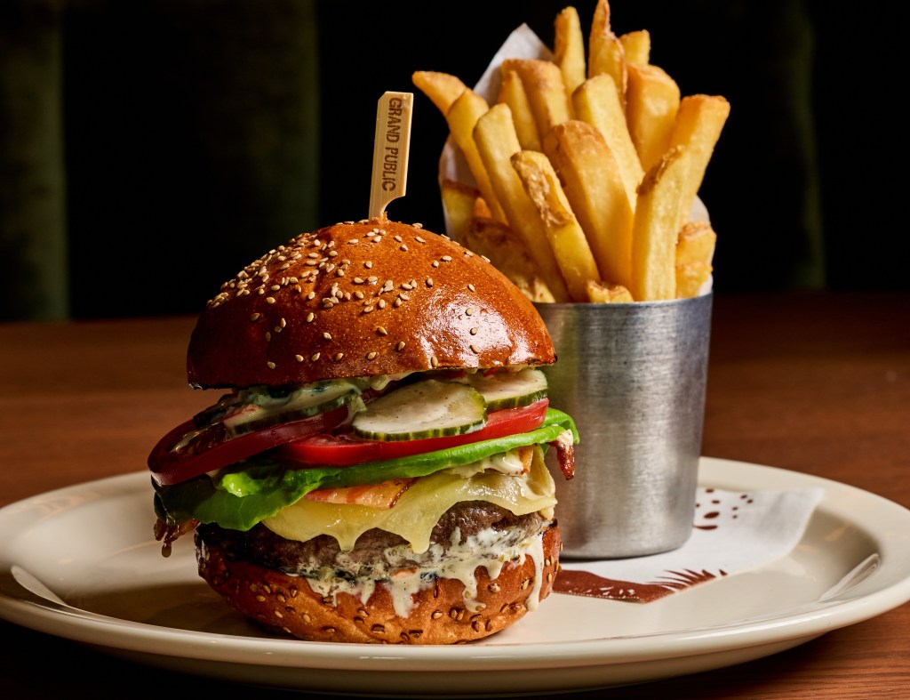 a cheeseburger with vegetable toppings next to a container of French fries on a wood table