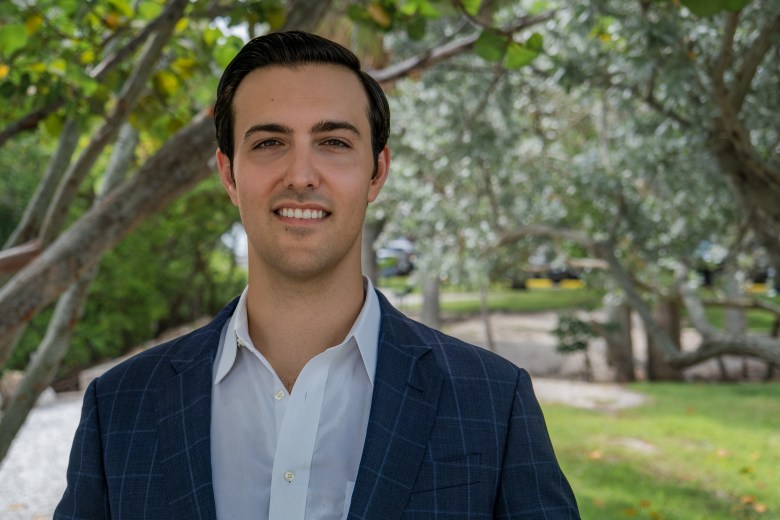 A professional outdoor campaign headshot of Garrett Greco smiling directly at the camera. The individual has short, neatly styled dark hair and is wearing a dark blue windowpane suit jacket over a crisp white button-down shirt with an open collar. They are standing against a softly blurred background featuring lush green trees, foliage, and a grassy park setting on a bright day.