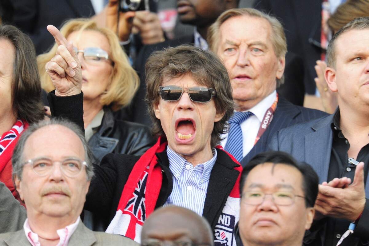 English musician Mick Jagger (C), flanked by former German football federation president and vice-president of the UEFA Gerhard Mayer-Vorfelder (background R),  cheers in the stands at the 2010 World Cup round of 16 football match England versus Germany on June 27, 2010 at Free State Stadium in Mangaung/Bloemfontein. NO PUSH TO MOBILE / MOBILE USE SOLELY WITHIN EDITORIAL ARTICLE -    AFP PHOTO / JOHN MACDOUGALL (Photo by JOHN MACDOUGALL / AFP via Getty Images)