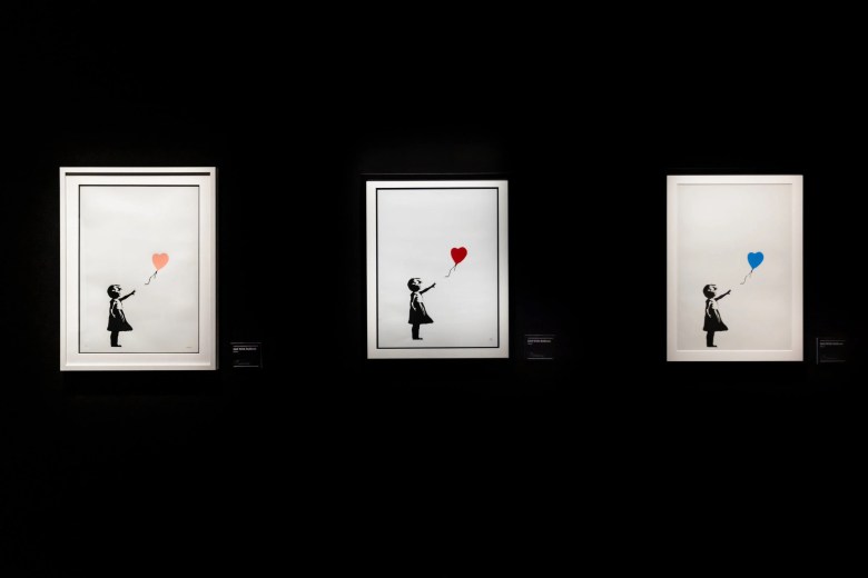A photograph by Kyle Flubacker displaying a triptych of three rare color variations of Banksy's iconic "Girl with Balloon" artwork, presented against a stark black background. The image features three identical white rectangular frames hung side-by-side. Each frame contains the famous black stenciled silhouette of a young child reaching upward toward a heart-shaped balloon blowing away in the wind. The balloon in the left frame is a pale pink, the center frame features a traditional bright red balloon, and the right frame displays a bright blue balloon.