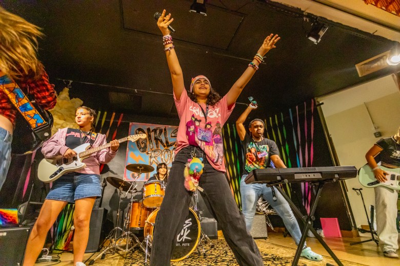 A vibrant live music photograph of a youth band performing on stage during Girls Rock Camp St. Pete on July 18, 2025. In the center, a musician stands triumphantly with both arms raised high in the air, holding drumsticks. They are wearing a pink graphic t-shirt, dark pants with a colorful fuzzy plush accessory attached to the waist, and multiple bracelets. To the left, a band member plays a white electric guitar, dressed in a layered purple and dark top with blue denim shorts. Behind them, another musician is seated at a wooden drum kit featuring the "Girls Rock St. Pete" logo on the kick drum. To the right, an individual plays a black keyboard on a stand, wearing a dark graphic t-shirt and light blue jeans. Partially visible band members play electric guitars on the far left and right edges of the frame. The stage is set with a colorful striped backdrop and illuminated by overhead spotlights from a dark ceiling.