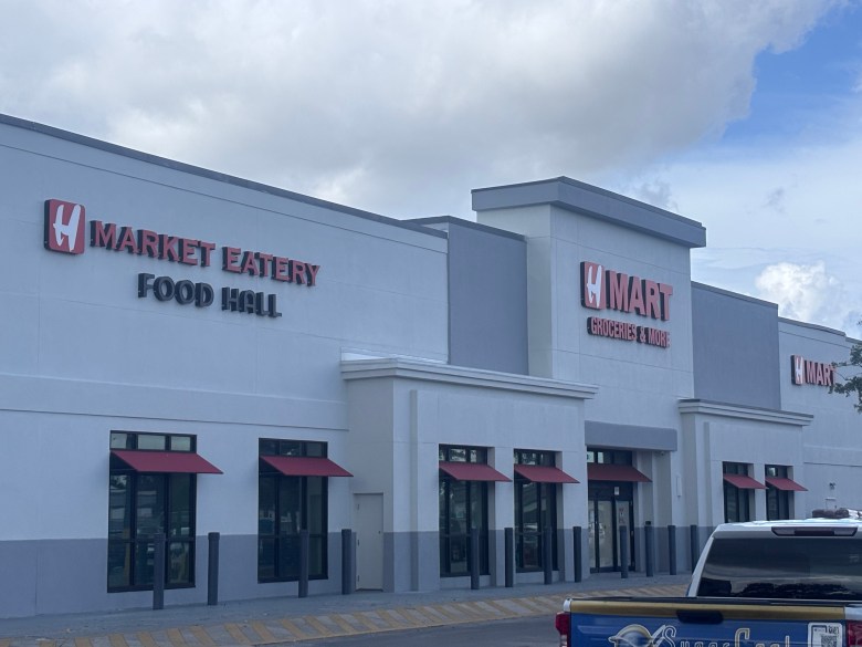An exterior photograph showing the storefront of the H Mart supermarket in Tampa, Florida, on July 20, 2026. The wide shot captures a modern, light grey and white building with dark grey accents and small red window awnings under a partly cloudy sky. On the left section of the building, bold lettering reads "H MARKET EATERY FOOD HALL," while the central entrance features the main "H MART" logo. In the lower right foreground, the tailgate of a dark blue pickup truck is partially visible in the parking lot.