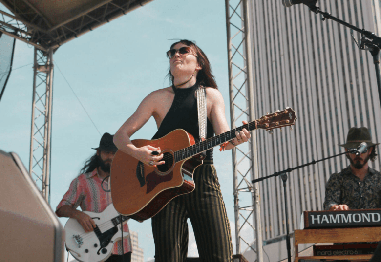A live performance photograph by Kasey Makal showing Hannah Harber performing on stage at the Gasparilla Music Festival in Kiley Garden in Tampa, Florida, on March 23, 2019. Positioned center stage, the lead performer stands holding and playing an acoustic guitar while looking upward with a passionate expression. They are wearing dark sunglasses, a black halter neck tank top, a black choker necklace, and dark vertically striped pants. In the background under bright daylight, two band members perform alongside: a bass player on the left in a pink and white striped shirt playing a white electric bass, and a keyboardist on the right wearing a hat and patterned shirt at a Hammond organ setup. Metal stage trusses and a tall building facade frame the background under a clear sky.