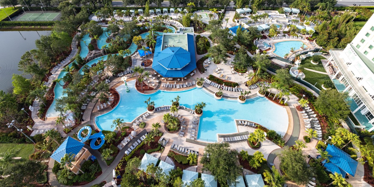 Hilton Orlando - Pool Aerial view of a large resort pool area with multiple swimming pools, water slides, lounge chairs, cabanas, and surrounding greenery.