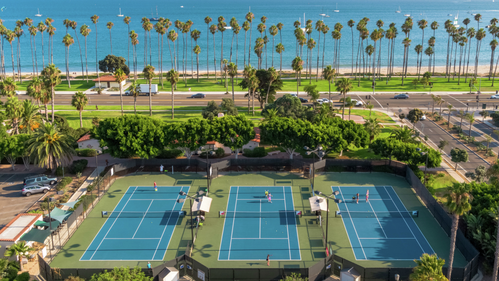 Three pickleball courts at the Hilton Santa Barbara Beachfront Resort with players, surrounded by trees and a road, with tall palm trees and the ocean in the background.