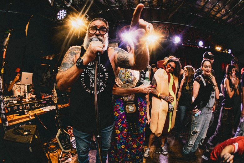 A wide-angle concert photograph by Dave Decker showing Crowbar venue owner Tom DeGeorge speaking to a crowd on stage during Hot Dog Party 20 at Crowbar in Ybor City, Florida, on July 3, 2026. Positioned center frame in a medium shot under bright stage lights, DeGeorge gestures upward with his left hand while speaking into a microphone. He features a full white beard and glasses, wearing a dark sleeveless graphic shirt displaying white text and jeans. Behind him on stage, several attendees and performers—including someone wearing a full-body hot dog costume and another wearing a wrestling championship belt—stand in front of DJ equipment against dark stage backdrop lighting.