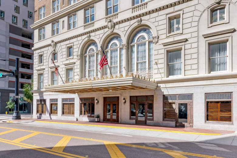 A eye-level exterior photograph showing the Cass Street entrance canopy and historic facade of Hotel Flor Tampa Downtown (formerly the Floridan Palace Hotel) in Tampa, Florida. Captured from across the street, the light beige stone building features grand arched second-story windows framed by ornamental reliefs, an American flag mounted on the right pillar, and a gold-trimmed entrance canopy extending over the wide sidewalk. At street level, dark wood-framed windows with louvered blinds line the ground floor leading to the double-door glass main entry, set along a street marked with bright yellow street markings.