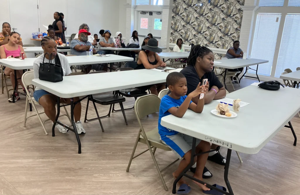 Attendees of a public meeting sit in rows of tables. 