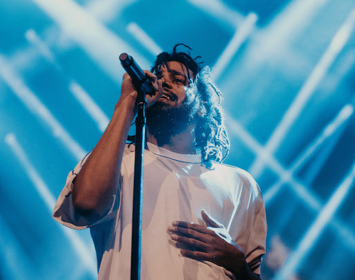 A live music photograph of hip-hop artist J. Cole performing on stage at Amalie Arena in Tampa, Florida, on August 11, 2018. Framed from the chest up, the musician leans in to sing into a microphone mounted on a straight stand, with their other hand resting flat against their chest. They have long braided hair and a beard, and are wearing a plain, light-colored short-sleeve t-shirt. The background features dynamic, vibrant blue stage lighting with striking, intersecting diagonal white light beams crossing behind the performer.