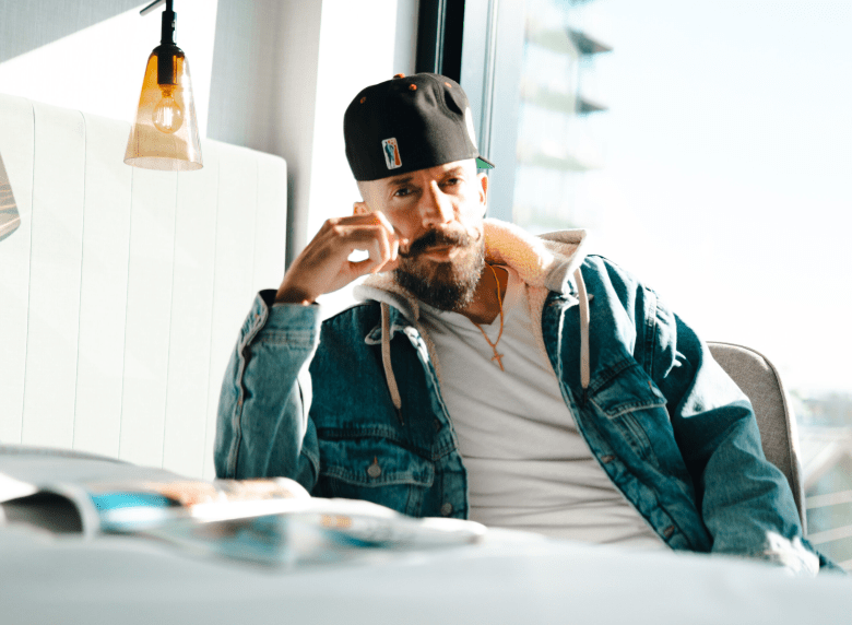 A brightly lit promotional portrait of Miami DJ Jesse Perez seated indoors next to a sunlit window. The musician is framed from the chest up, looking directly at the camera with one hand raised, resting lightly near their prominent, styled curled mustache and full dark beard. They are wearing a black baseball cap featuring a small square logo, a classic blue denim jacket layered over a grey hoodie, a plain white t-shirt, and a thin gold chain necklace. In the softly blurred foreground, an open magazine rests on a white table. The background features bright natural daylight streaming in, illuminating a clear glass hanging pendant light with an Edison-style bulb visible on the left side of the frame.