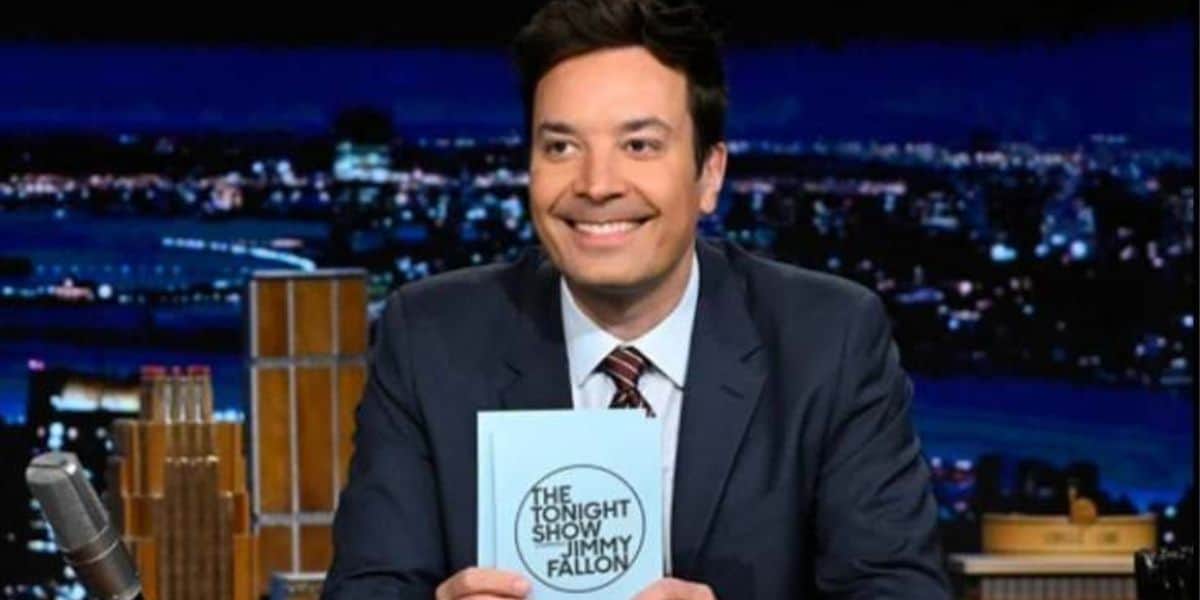 A man in a suit and tie sits at a desk during a nighttime TV show, smiling and holding up a book titled "Jimmy Fallon: The Tonight Show" against a backdrop of a city skyline. A microphone is visible on the desk.