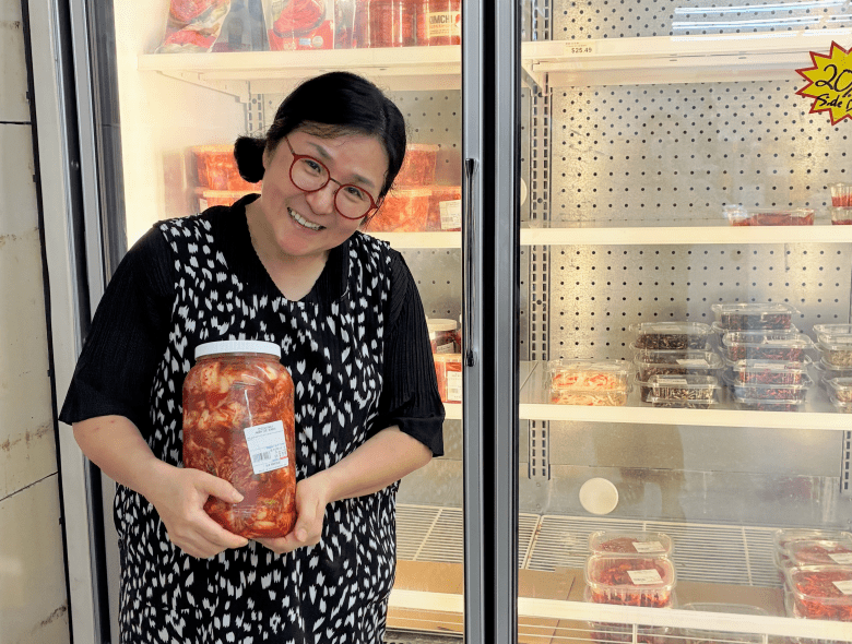A portrait photo by Annalise Mabe of Jin Cha holding a large glass jar of homemade kimchi at Kim Bros. Korean market in Tampa, Florida, on July 20, 2026. Positioned center frame, the individual smiles warmly at the camera, wearing red round-framed glasses, a dark patterned black-and-white dress over a dark short-sleeved top, and dark hair styled in low pigtails. They are cradling the large transparent jar of kimchi with both hands in front of open commercial refrigerator cases filled with stacked food containers, packaged side dishes (banchan), and a yellow starburst price tag on a wire shelf in the background.