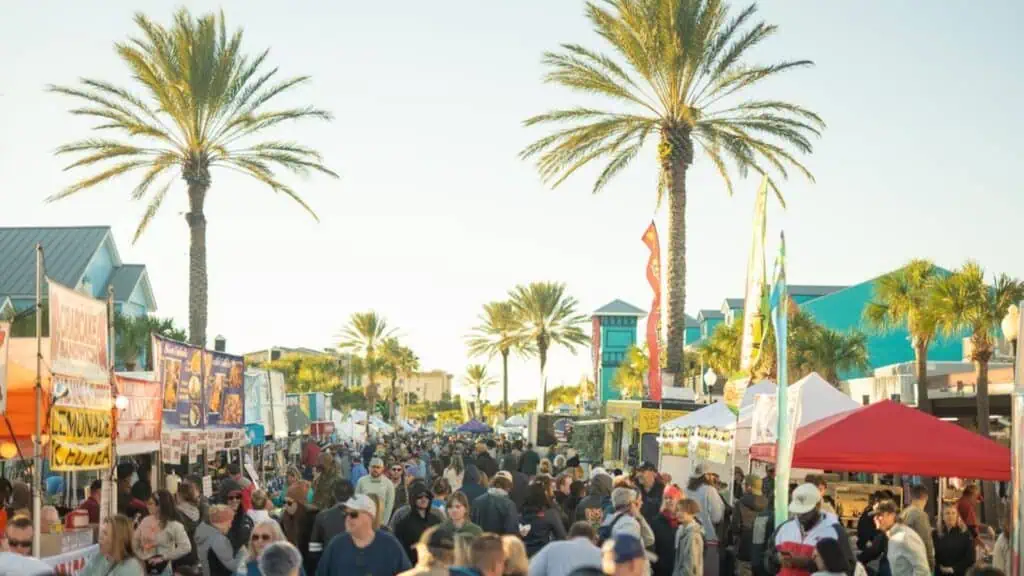 a large group of people are shopping and enjoy food from a variety of vendors dooring an open-air seafood festival in the John's Pass area of Pinellas County. Tall palm trees line the sidewalks. It's a bright and sunny day.