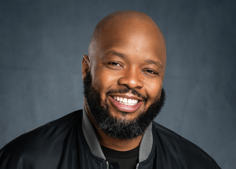 A promotional portrait of comedian Kevin Fredericks, aka KevOnStage, smiling warmly directly at the camera. Framed from the chest up, the individual is bald with a full, dark beard and mustache. They are wearing a black bomber-style jacket with a grey ribbed collar, layered over a plain black t-shirt. The background features a mottled, textured dark grey-blue backdrop.
