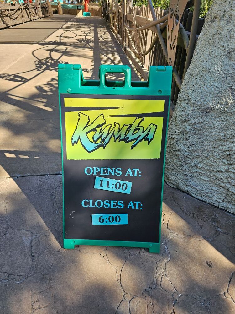 A green-and-black A-frame sign on a stone pathway showing the operating hours, reading "opens at 11:00 and closes at 6:00."
