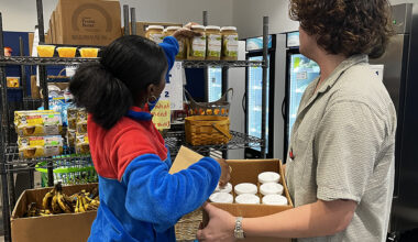 A woman in a blue and red fleece jacket reaches up to arrange jars of applesauce on a metal shelf, while a person beside her holds a cardboard box filled with glass jars. Surrounding shelves and boxes contain peanut butter, bananas, juice boxes, and commercial refrigerators.