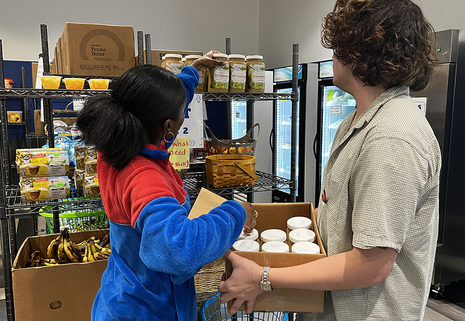 A woman in a blue and red fleece jacket reaches up to arrange jars of applesauce on a metal shelf, while a person beside her holds a cardboard box filled with glass jars. Surrounding shelves and boxes contain peanut butter, bananas, juice boxes, and commercial refrigerators.