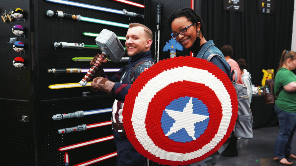 A photograph of two attendees posing with fan-made props at Brick Fan Fest. In the center, two individuals smile together—one wearing a Captain America suit and holding Thor’s hammer (Mjölnir), and another standing behind a large, circular Captain America shield constructed entirely out of red, white, and blue LEGO bricks. To the left, a black wall display holds various glowing and non-glowing toy lightsabers with colorful hilts. In the background, other convention attendees move through an open event hall set up with display tables and black curtains.