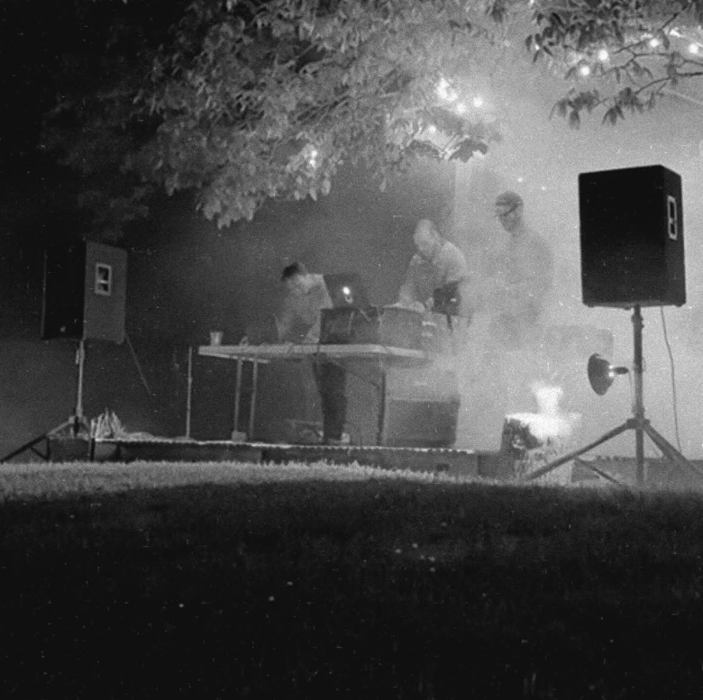 A black-and-white photograph capturing an atmospheric, smoky outdoor live music performance by the band LMH. Framed through a wide shot, three band members are positioned behind a long folding table set up on a small grass stage, surrounded by equipment. The musicians appear as soft, slightly obscured figures amidst thick theatrical fog, with two standing on the right and one leaning over equipment on the left. The stage is flanked by two large, black PA speakers on tall tripod stands. Strung overhead are glowing warm patio bulb lights, partially obscured by the leaves of a large overhanging tree. The foreground is dominated by a dark, grassy lawn, creating a distinct contrast with the illuminated, hazy stage setup.