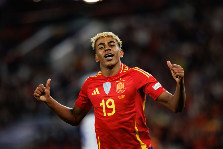 A sports photograph of soccer player Lamine Yamal celebrating a goal during the UEFA Nations League 2025 semifinal game between Spain and France at MHPArena in Stuttgart, Germany, on June 5, 2025. The athlete is framed from the waist up against a deeply blurred, dark stadium background, smiling widely and looking forward with both hands raised, giving a double thumbs-up gesture. They have short, textured dark hair with frosted blonde tips and are wearing a bright red Spain national team jersey featuring yellow shoulder stripes, the team crest, a UEFA patch, and the number 19 printed in yellow on the center of the chest.