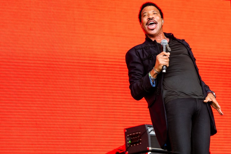 A live music photograph by Anthony Mooney of musician Lionel Richie performing on stage at the Glastonbury Festival in Somerset, United Kingdom, on June 28, 2015. The artist is captured from the thighs up, leaning slightly back and smiling widely while singing into a microphone held in their right hand. They are dressed in an all-black ensemble, including a dark t-shirt, dark fitted trousers, and a long black overshirt or light jacket. The performer stands in front of a massive, vibrant orange LED screen that serves as a bold, solid background, with a small black stage monitor partially visible at their feet.