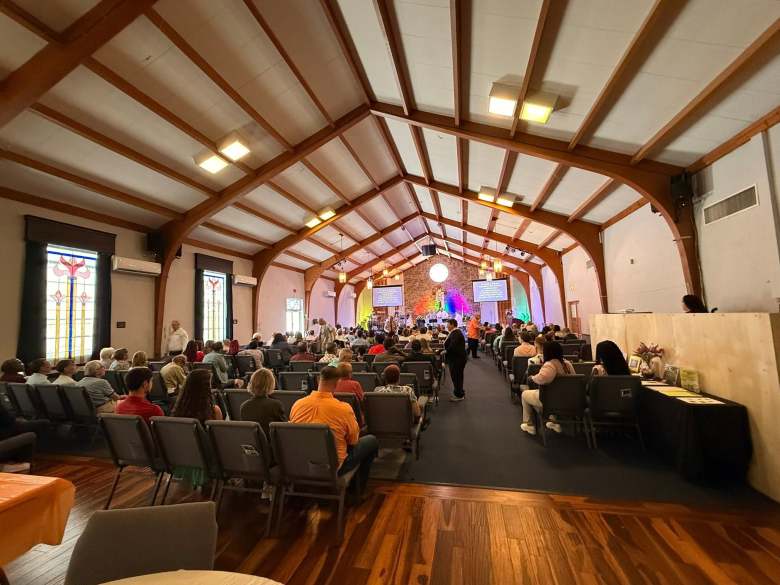 A wide-angle interior photograph showing the sanctuary filled with congregants during a service at Metropolitan Community Church of Tampa (MCC Tampa) in Tampa, Florida, on April 5, 2026. Taken from the back of the worship hall facing forward, rows of grey padded chairs line both sides of a central carpeted aisle under a high vaulted ceiling with exposed wooden beams and rectangular light fixtures. On the left wall, tall stained-glass windows cast colorful light into the room, while congregants sit facing the front altar. The altar area features a natural stone backdrop, a glowing circular display, vibrant rainbow accent lighting, and two elevated projection screens displaying service text.