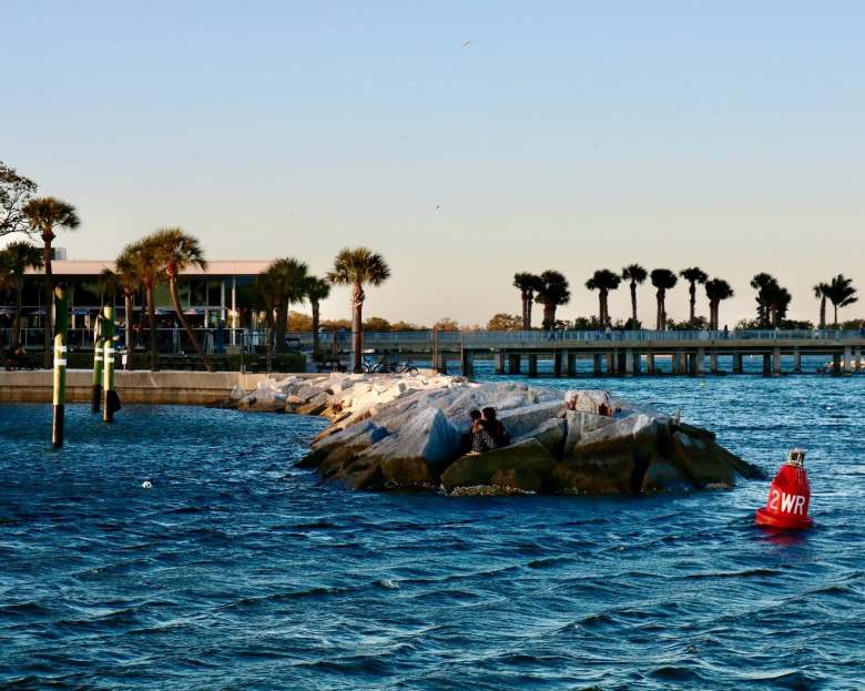 A sunny, wide landscape photograph taken from the waters surrounding the St. Pete Pier in St. Petersburg, Florida, capturing a scenic coastal view. In the midground, a long, low breakwater made of light-colored, jagged rocks juts out into the calm, deep blue water, with two figures sitting together on the rocks near the center. To the right, a bright red buoy labeled "2WR" floats on the water's surface. In the background, green palm trees and coastal buildings line the shore under a clear, pale blue sky, with a long pedestrian pier structure extending horizontally across the right side.